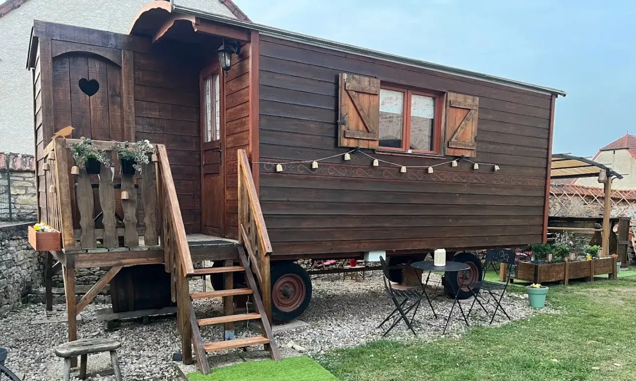 Charmante roulotte en bois, parfaite pour un camping de la ferme. Escaliers en bois, jardini&egrave;res fleuries et petite table ext&eacute;rieure invitent &agrave; la d&eacute;tente.