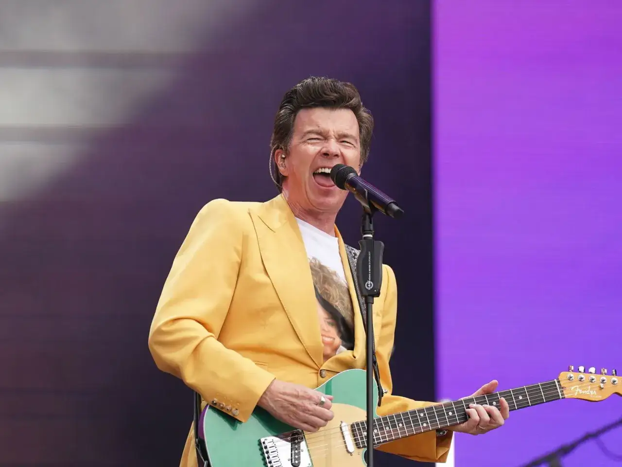Rick Astley, jung und energiegeladen, rockt die B&uuml;hne in einem gelben Sakko und spielt Gitarre.