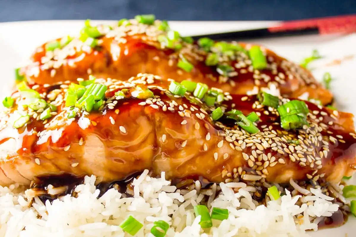 Två saftiga filéer av teriyaki lax, toppade med sesamfrön och grön lök, serveras över en bädd av fluffigt ris.