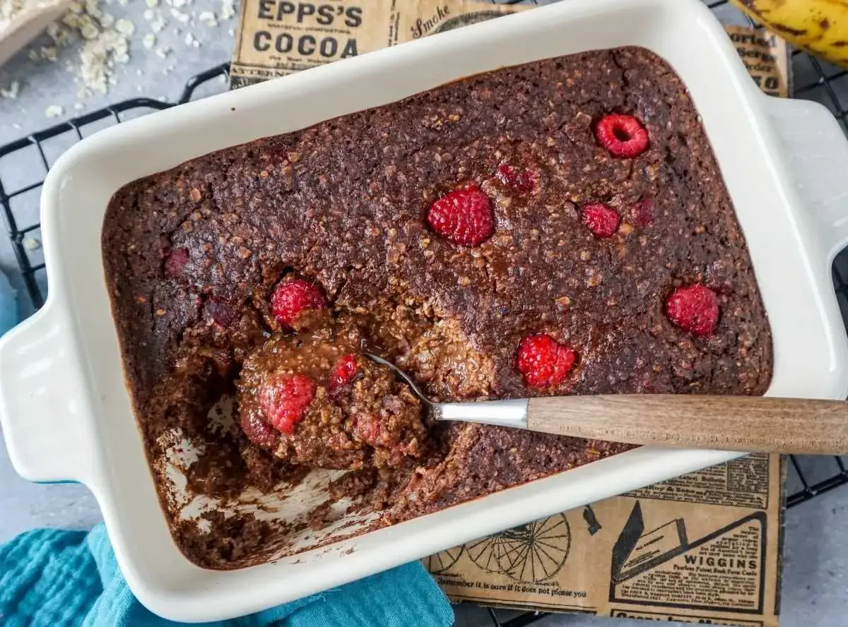 Schokoladiger baked oats mit Himbeeren in einer wei&szlig;en Auflaufform, eine Portion wird mit einem L&ouml;ffel herausgenommen.