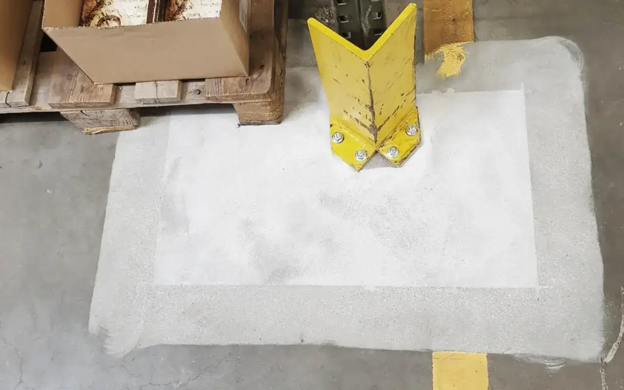 Naprawa popękanej posadzki betonowej: biała masa wypełnia ubytek, obok żółty odbojnik i paleta z kartonami.
