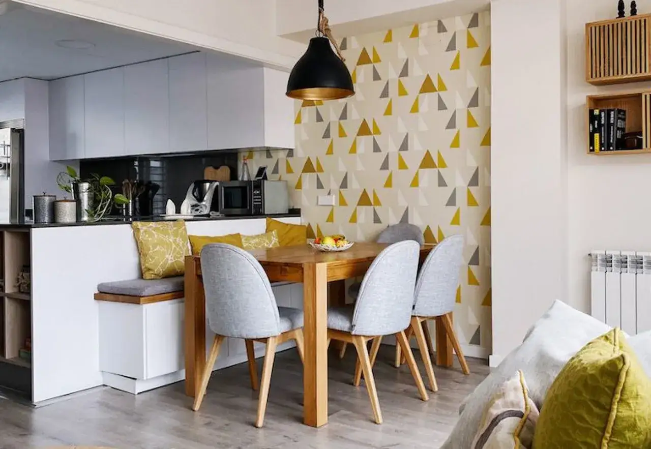 Comedor moderno con **cocinas con papel pintado** de triángulos amarillos y grises. Mesa de madera, sillas grises y banco con cojines.