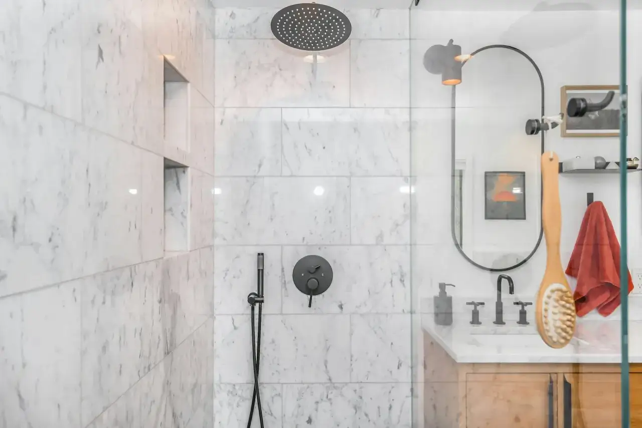 Ducha empotrada con grifo negro y rociador superior. La pared de m&aacute;rmol blanco tiene nichos. El espejo ovalado y el lavabo de madera completan el espacio.