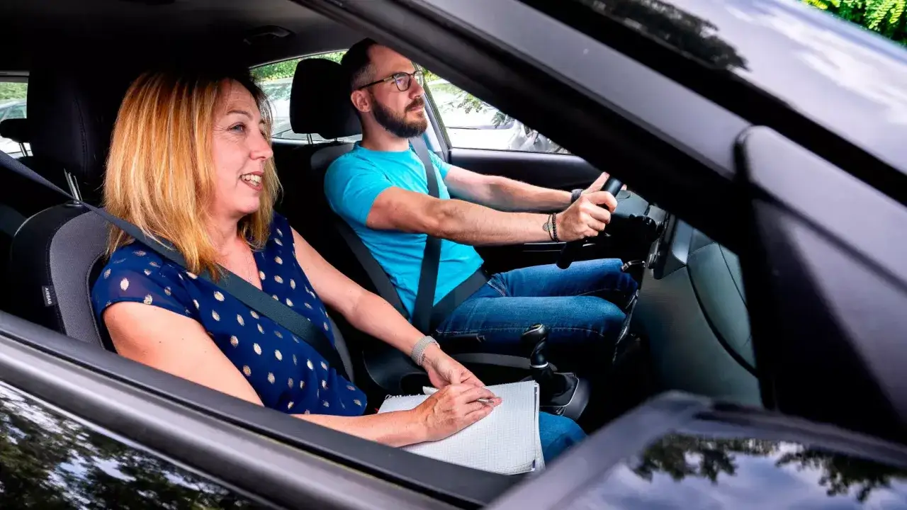 Une femme souriante, assise à côté du conducteur, tient un carnet. L'homme au volant semble concentré, peut-être lors d'un **examen du permis de conduire**.