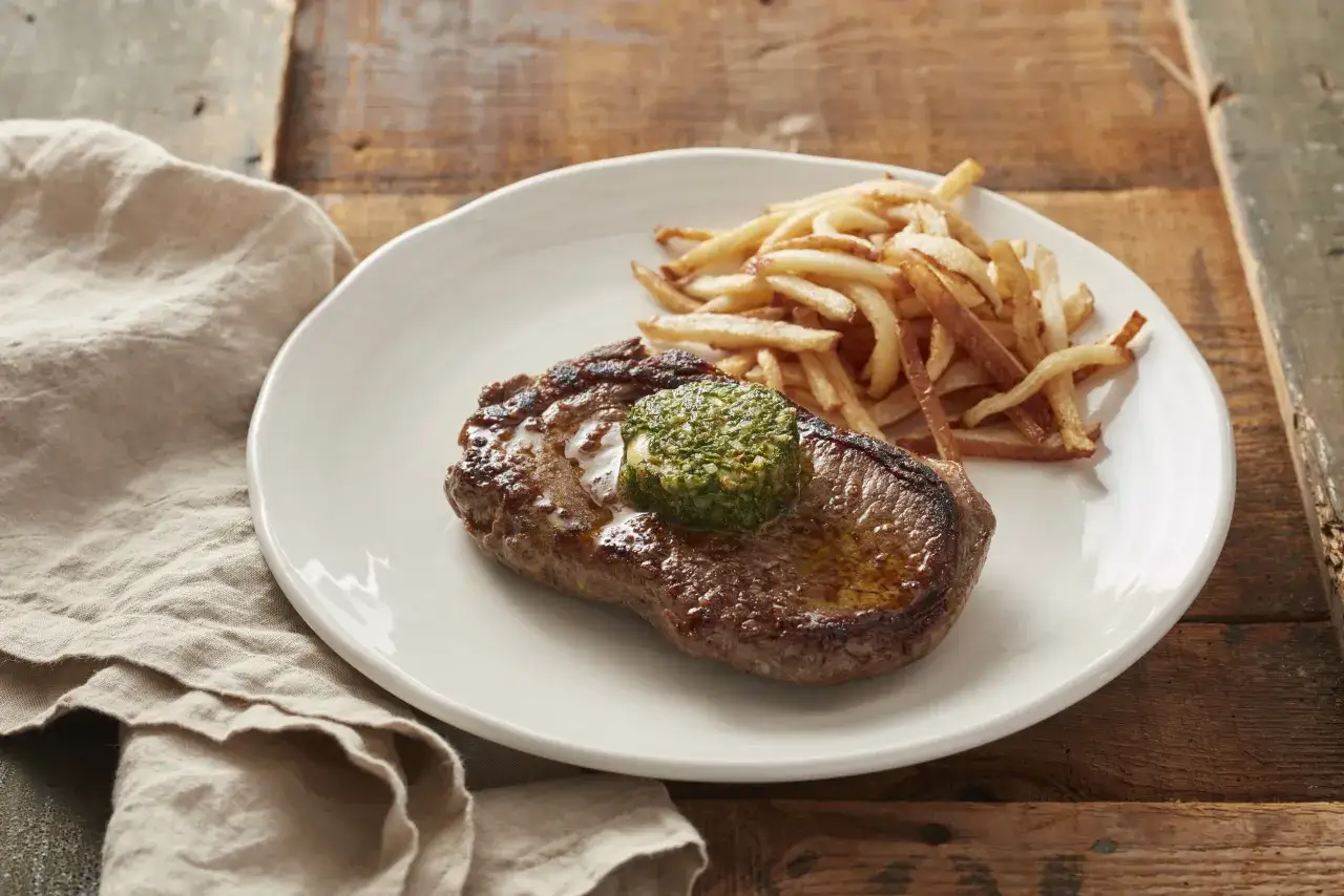 En saftig biff med caf&eacute; de Paris-sm&ouml;r och pommes frites p&aring; en vit tallrik, serverad p&aring; ett rustikt tr&auml;bord.