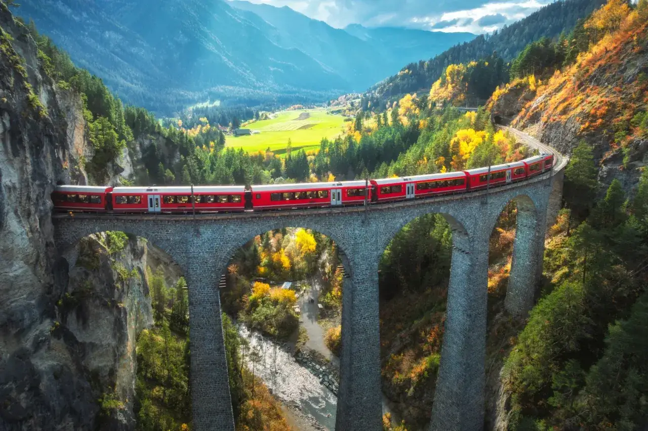 Roter Zug überquert eine Steinbrücke in den Alpen. Die Herbstfarben leuchten, während die Fahrt nach Rom beginnt.