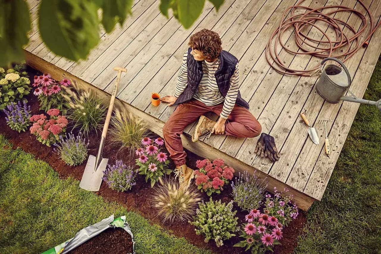 Person sitter på altanen och planerar en rabatt med blommor, gräs och jord. Trädgårdsredskap, vattenkanna och slang ligger bredvid.