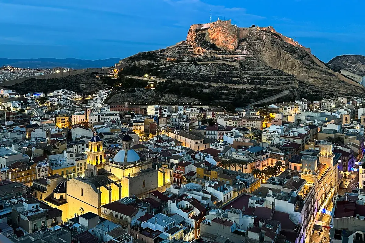 Alicante nocą, z zamkiem Santa Bárbara w tle. Widać oświetlone budynki i kościoły. Idealna pogoda w marcu na zwiedzanie.