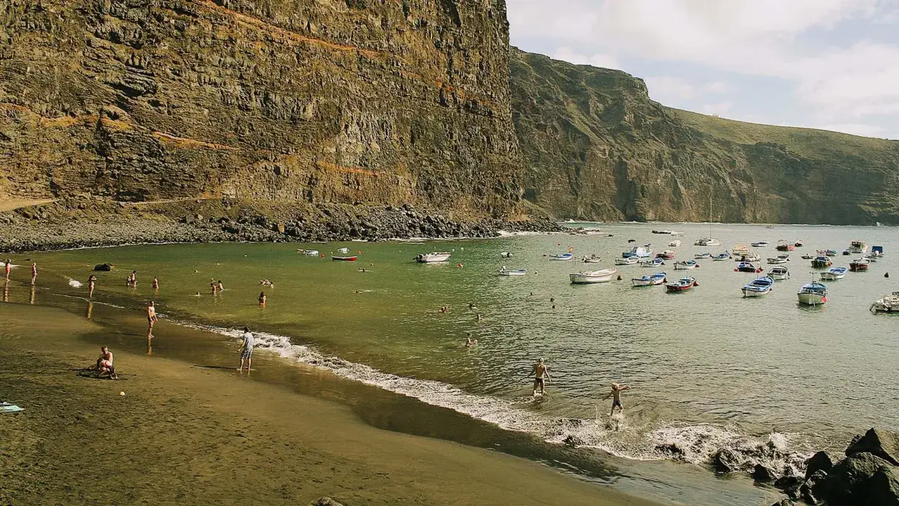 Gente disfruta del agua y barcos anclados en la playa de Guayedra, con imponentes acantilados de fondo.