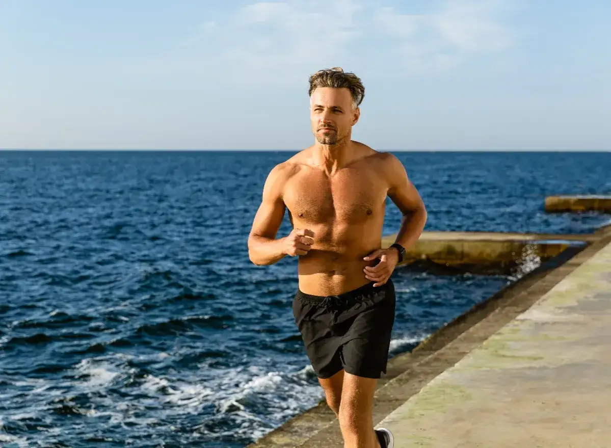 Ein normal trainierter Mann l&auml;uft am Meer entlang. Die Sonne scheint, das Meer ist blau.