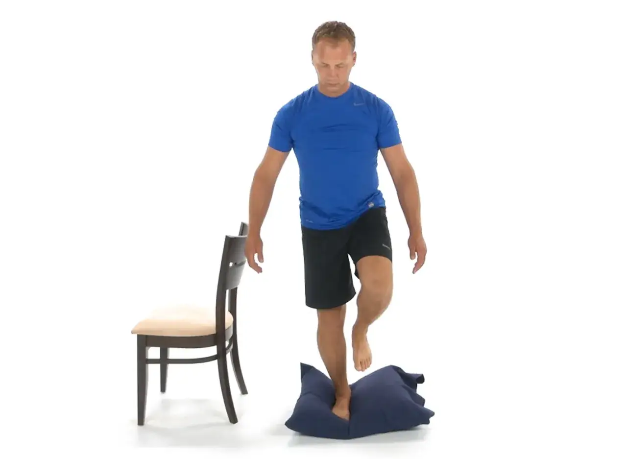 Uomo in piedi su un cuscino blu, con una gamba sollevata, per rafforzare le caviglie deboli. Accanto, una sedia per supporto.