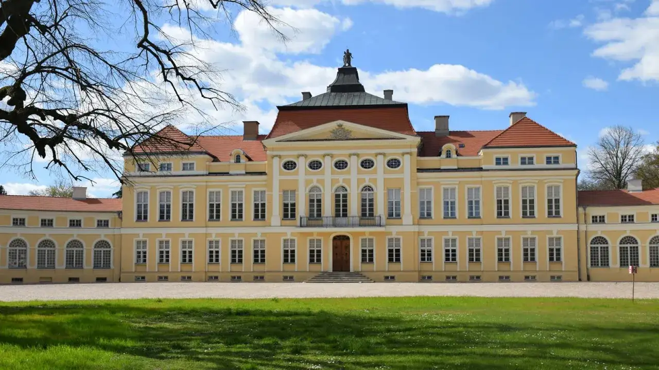 Piękny, żółty pałac w Wielkopolsce z czerwonym dachem i licznymi oknami, otoczony zielonym trawnikiem i drzewami.