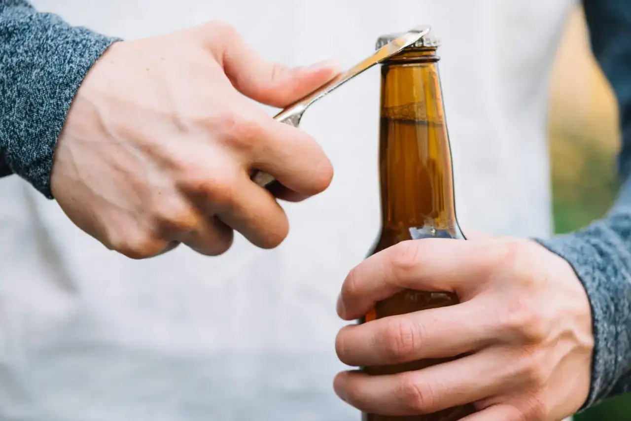 Main gauche tenant une bouteille, main droite utilisant un objet pour soulever le bouchon. Astuce pour savoir comment ouvrir une bouteille sans tire bouchon.