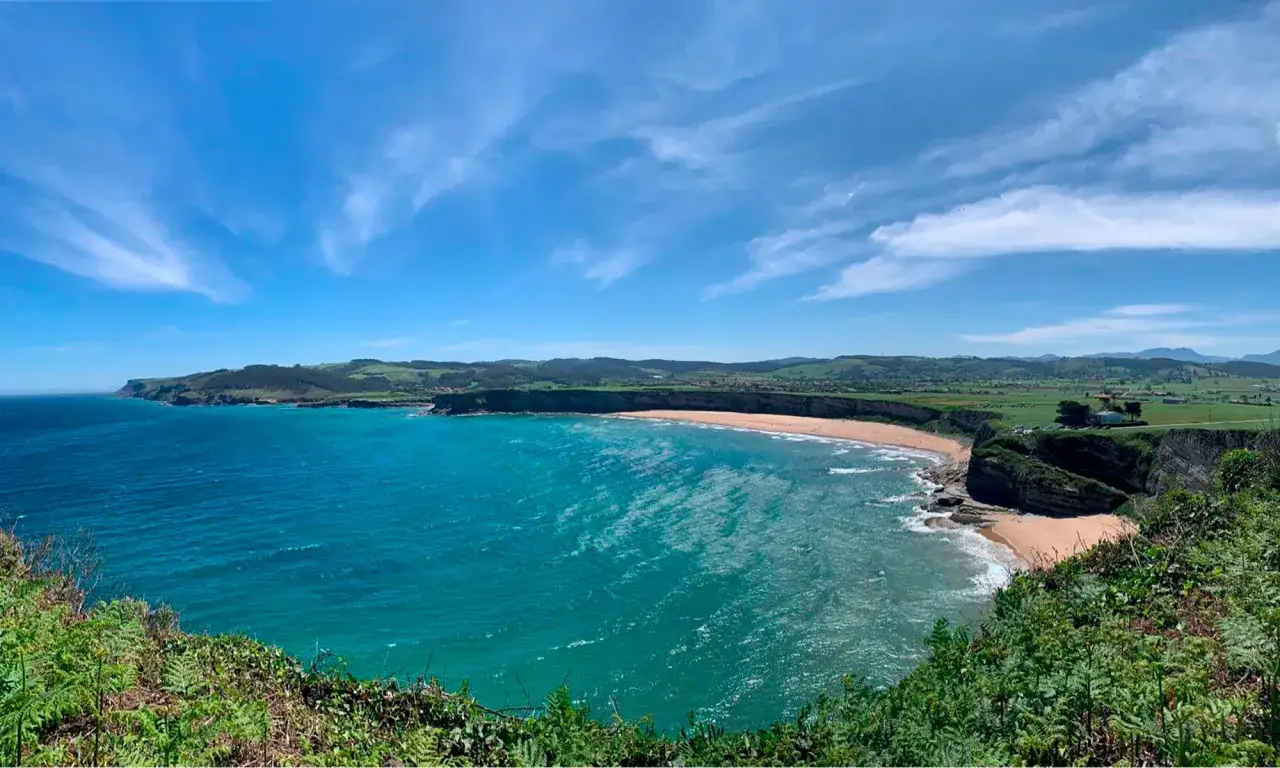 Costa verde y playa dorada, un rincón de curiosidades de Cantabria. El mar turquesa besa la arena bajo un cielo azul con nubes algodonosas.
