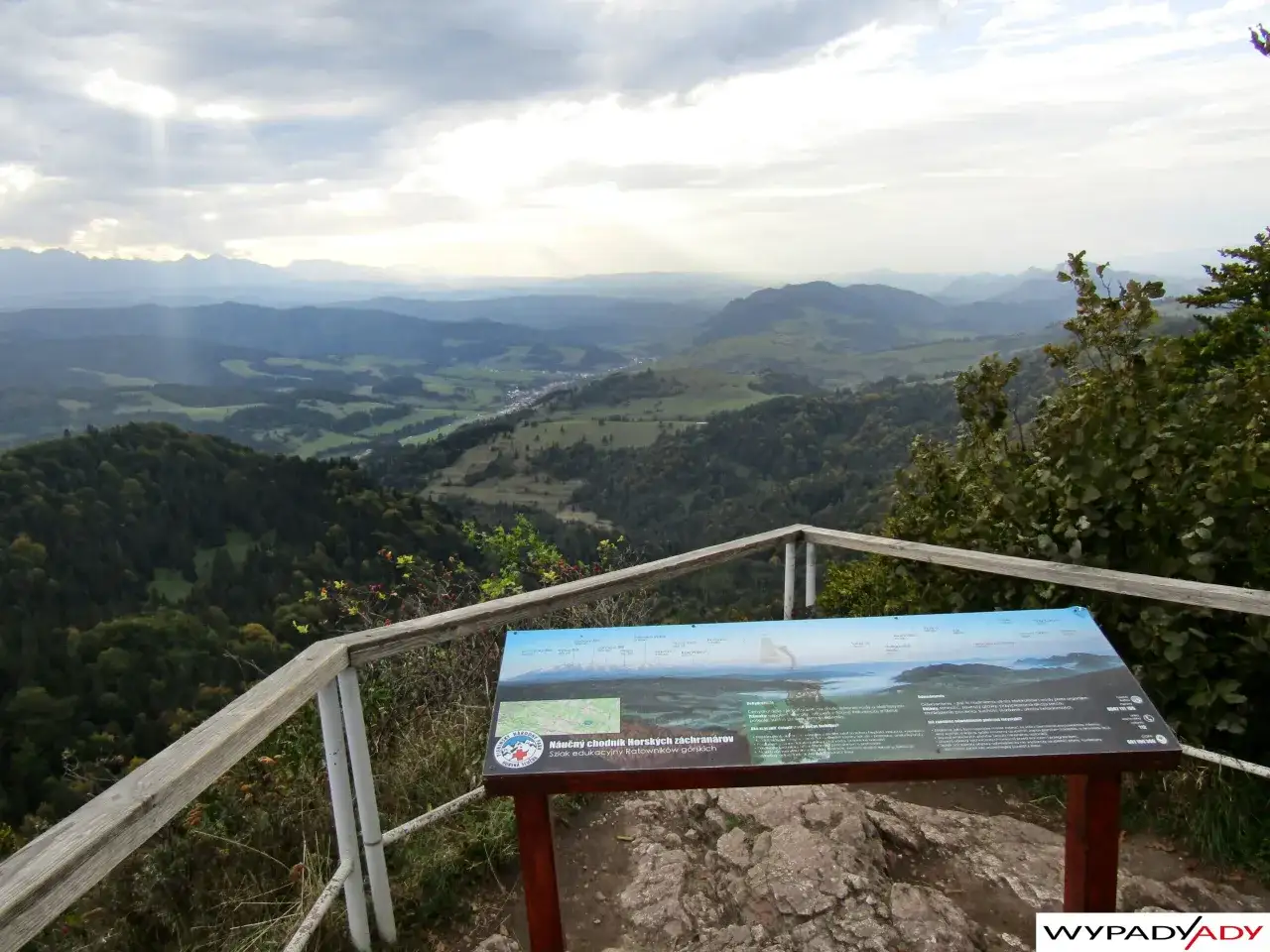 Widok na **wysokie Pieniny** z punktu widokowego. W oddali pasma górskie, chmury i promienie słońca. Na pierwszym planie tablica informacyjna.