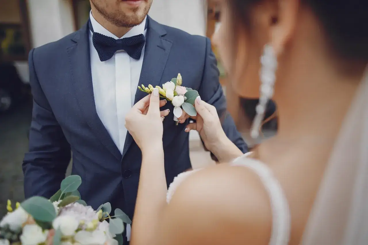 Panna młoda przypina butonierkę panu młodemu. Elegancka mucha dodaje mu uroku.