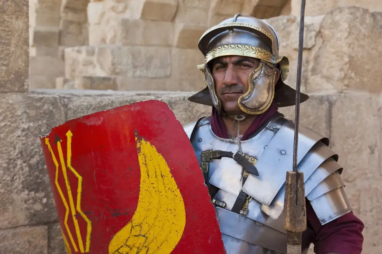 Rzymski legionista w zbroi i hełmie, z tarczą ozdobioną symbolem nawiązującym do Marsa, boga wojny z mitologii.