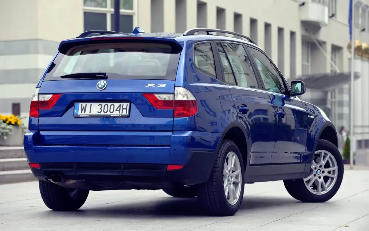 Niebieskie BMW X3 2005 z polskimi tablicami WI 3004H zaparkowane przed nowoczesnym budynkiem.