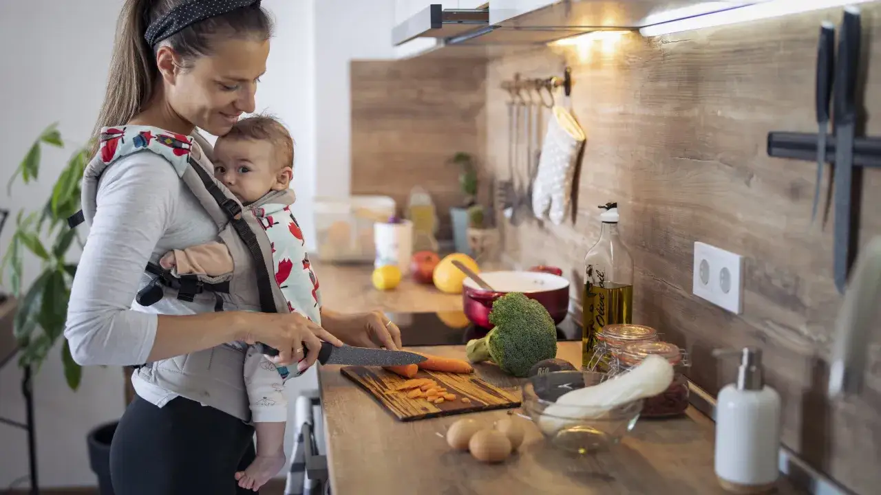 Mutter bereitet Essen zu, w&auml;hrend sie ihr Baby im Tragetuch h&auml;lt. Vielleicht genie&szlig;t sie danach eine Tasse rooibos tee stillzeit.