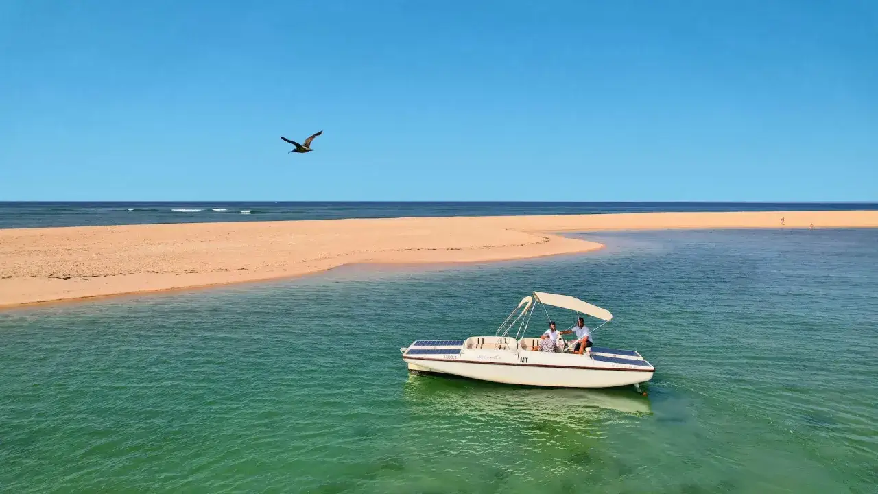 Ein Boot mit zwei Personen gleitet durch das türkisfarbene Wasser vor den Faro Inseln. Ein Vogel fliegt über der Sandbank.
