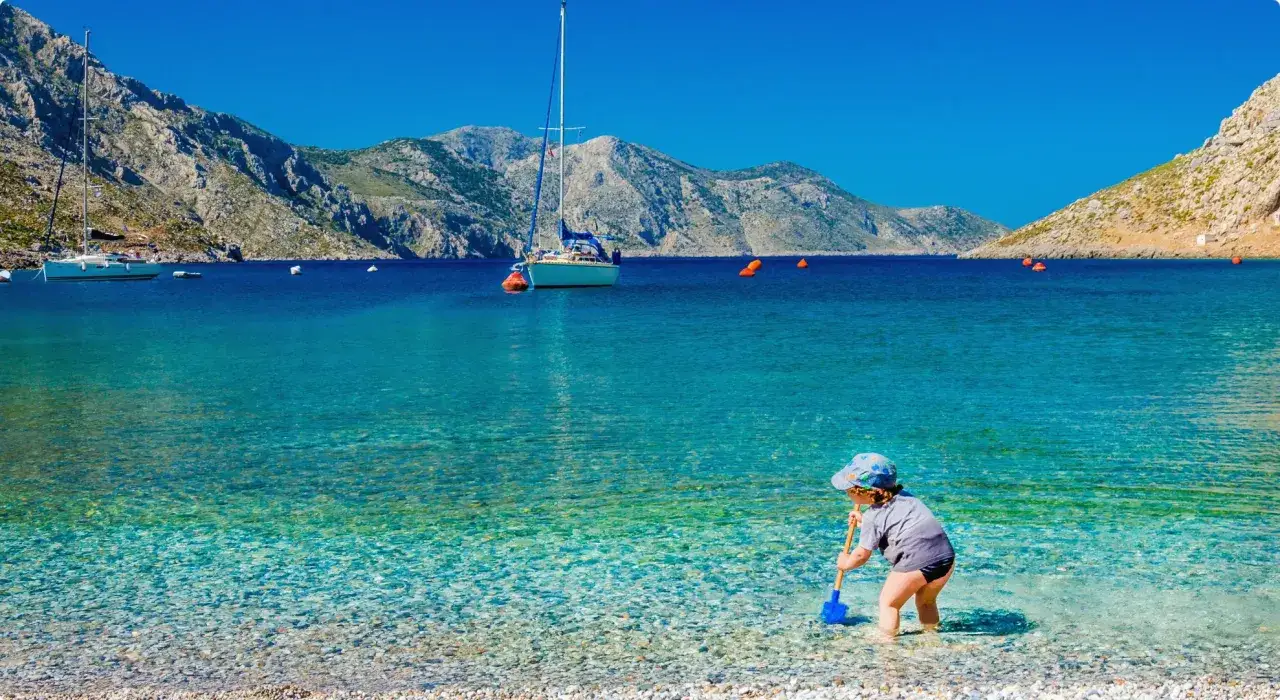 Małe dziecko z łopatką bawi się na kamienistej plaży. W tle błękitne morze, g&oacute;ry i żagl&oacute;wki. Cudowny dzień na Cyprze z dzieckiem.