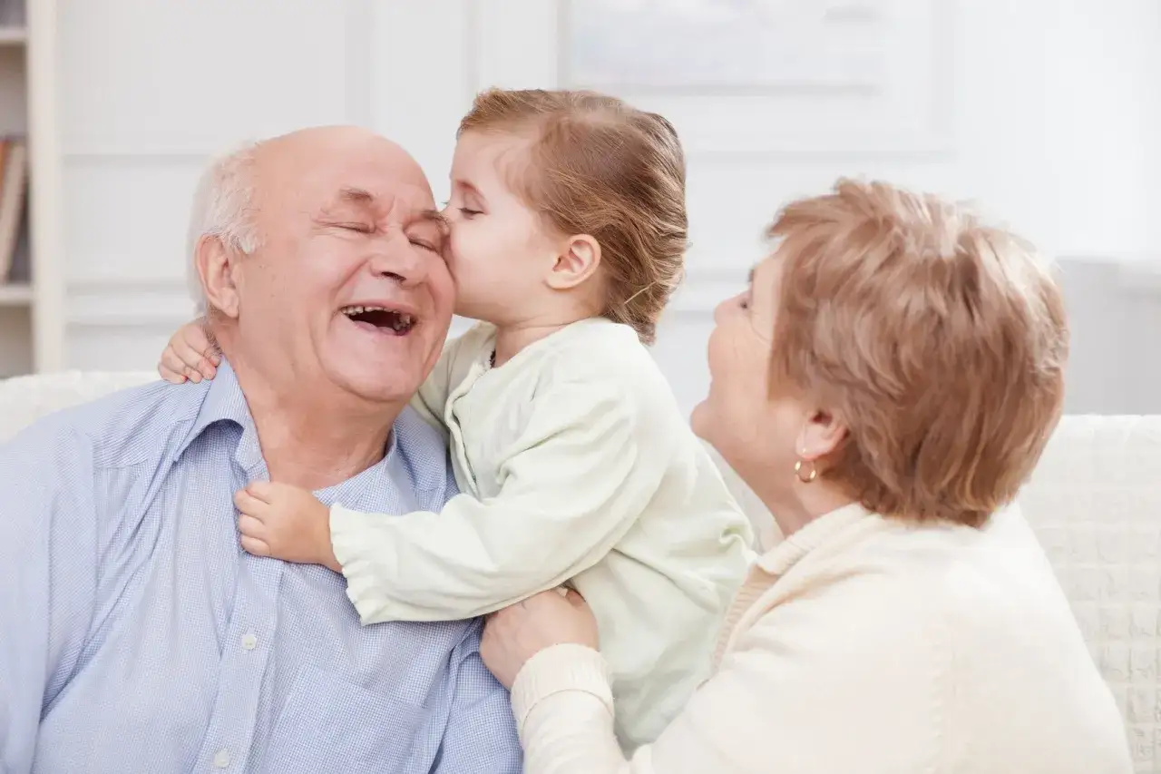 Un niño besa a su abuelo, quien ríe feliz. La abuela sonríe. Perfectas dedicatorias frases abuelos cortas para este momento.