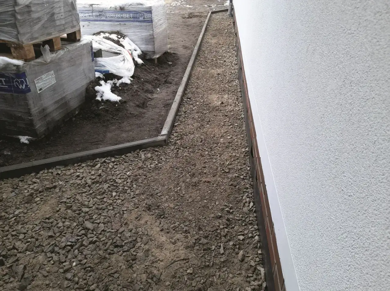 Wykonano opaskę wokół domu z tłucznia, obok ułożone są palety z materiałami budowlanymi.