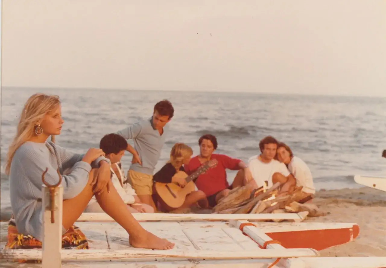 Giovani su una spiaggia, un gruppo di amici che ricorda i film estivi italiani, con una chitarra e il mare sullo sfondo.