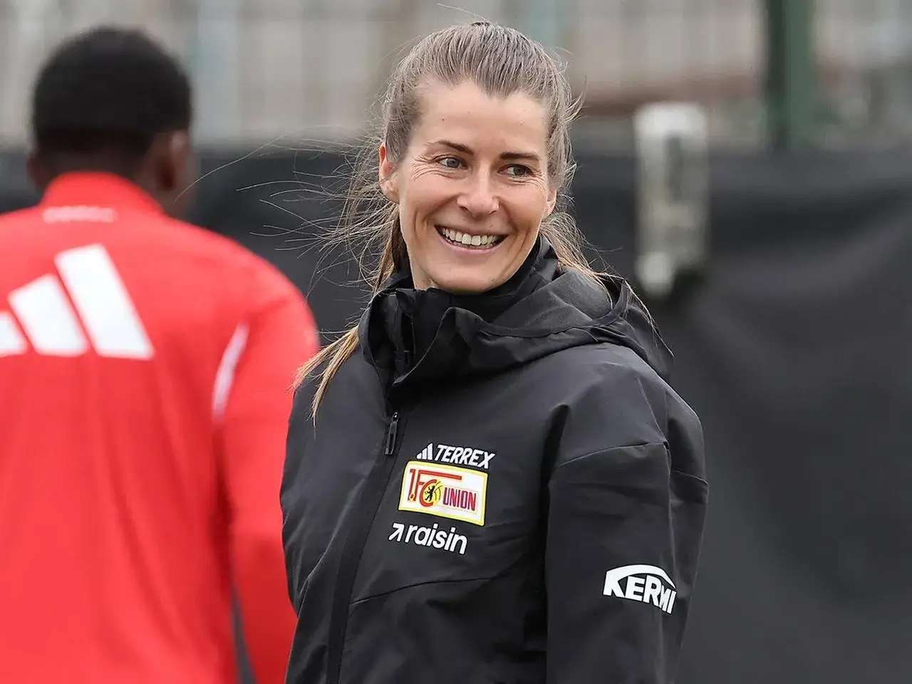 Marie-Louise Eta lacht fr&ouml;hlich auf dem Trainingsgel&auml;nde, tr&auml;gt eine schwarze Jacke mit dem Union Berlin Logo.