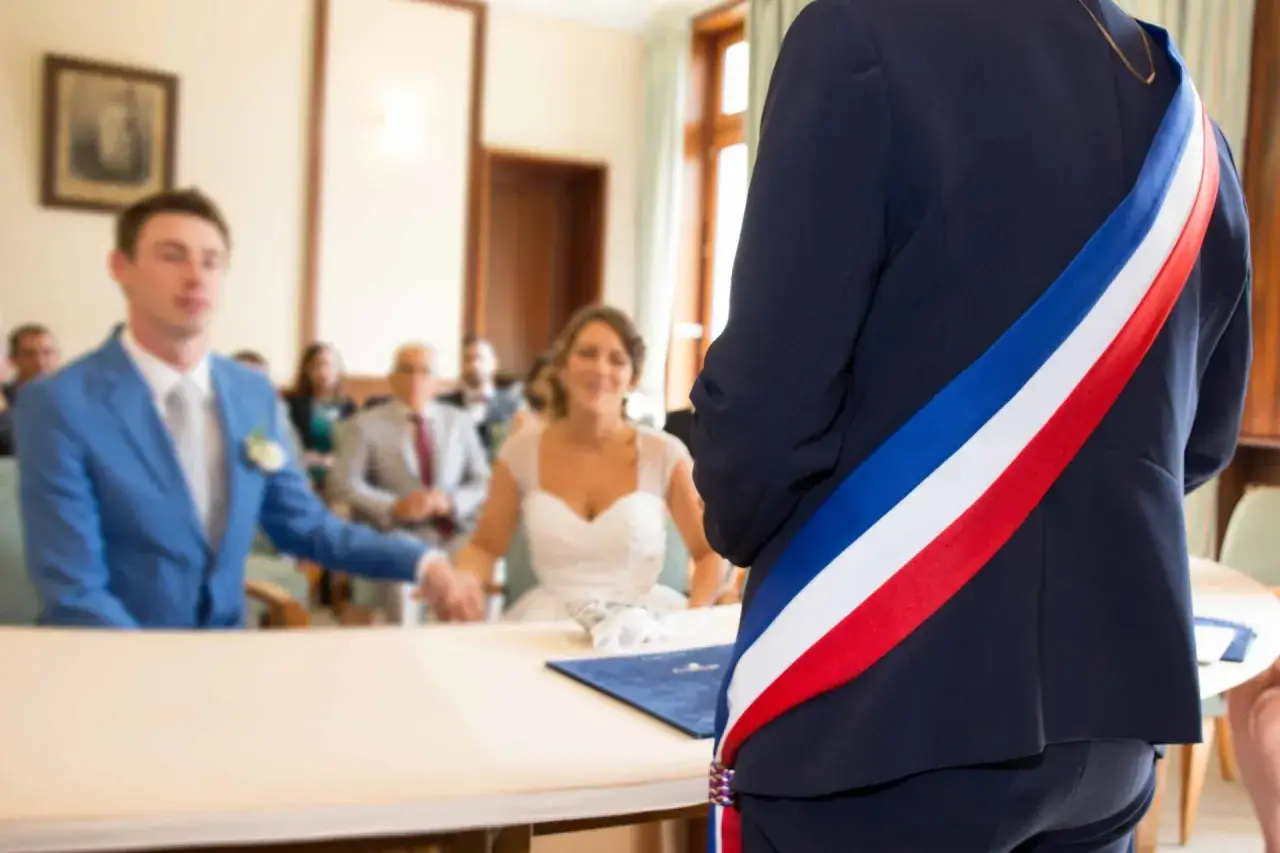 Cérémonie de mariage civil. Le maire, portant l'écharpe tricolore, officie. Le couple est assis, souriant. Le baptême civil prix est une démarche symbolique.