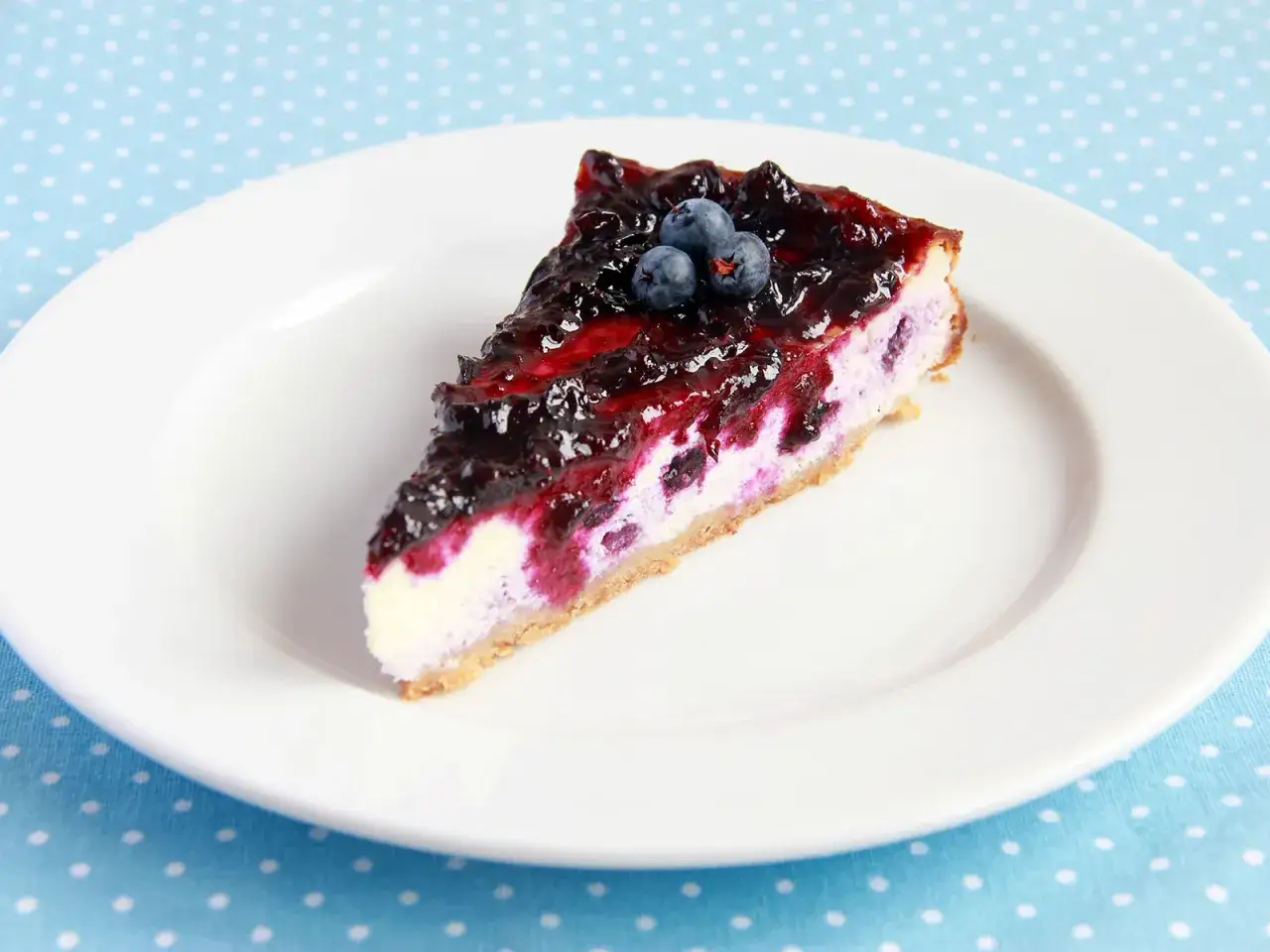 Ein Stück cremiger Blueberry Cheesecake mit Blaubeersauce und frischen Beeren, serviert auf einem weißen Teller.