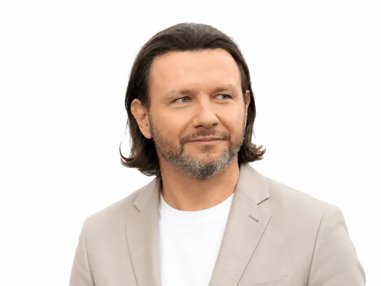 Mężczyzna z siwiejącą brodą i długimi włosami, ubrany w jasną marynarkę, patrzy w bok. Jego **majdan wzrost** sugeruje pewność siebie.