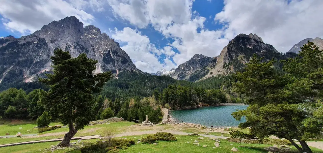 Paisaje alpino en el Vall de Boí Aigüestortes, con un lago azul turquesa, bosques de pinos y montañas nevadas bajo un cielo nublado.