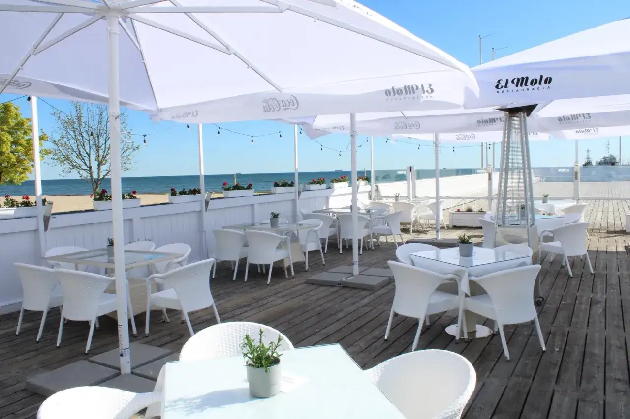 Letnie popołudnie w Trójmieście. Restauracja z widokiem na morze, białe meble i parasole tworzą idealne miejsce na relaks.