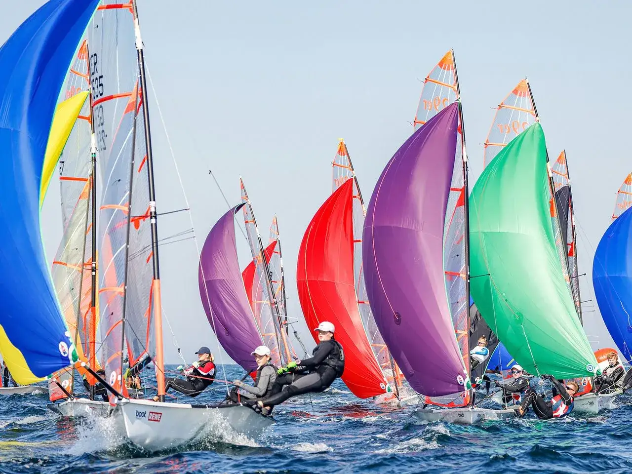 Bunte Segel dominieren das Bild beim Kieler Woche Segeln. Mehrere Boote mit Crews k&auml;mpfen auf dem Wasser um die Wette.