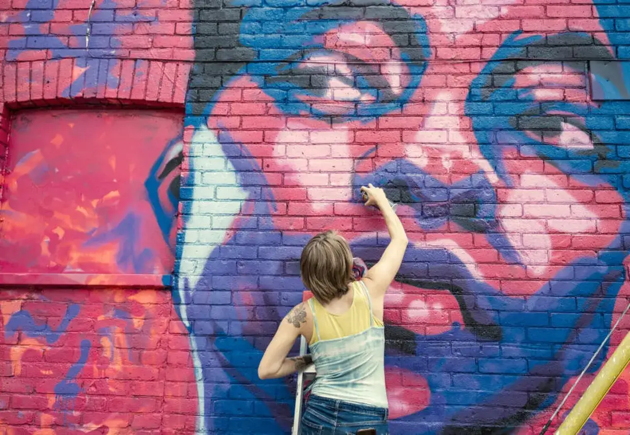 K&uuml;nstlerin spr&uuml;ht ein riesiges Portr&auml;t auf eine Ziegelwand. Ein Beispiel f&uuml;r **popular art**, das die Stadt versch&ouml;nert.