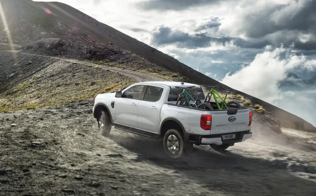Vit Ford Ranger laddhybrid med cyklar i flaket k&ouml;r p&aring; en grusv&auml;g i bergig terr&auml;ng.
