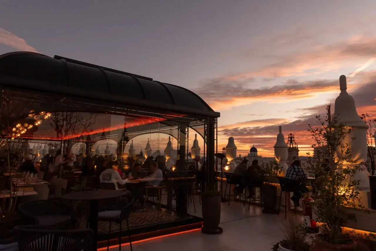 Atardecer en una terraza con vistas a la ciudad, bajo un cielo atenea. Gente cenando en un restaurante con cristaleras y luces cálidas.