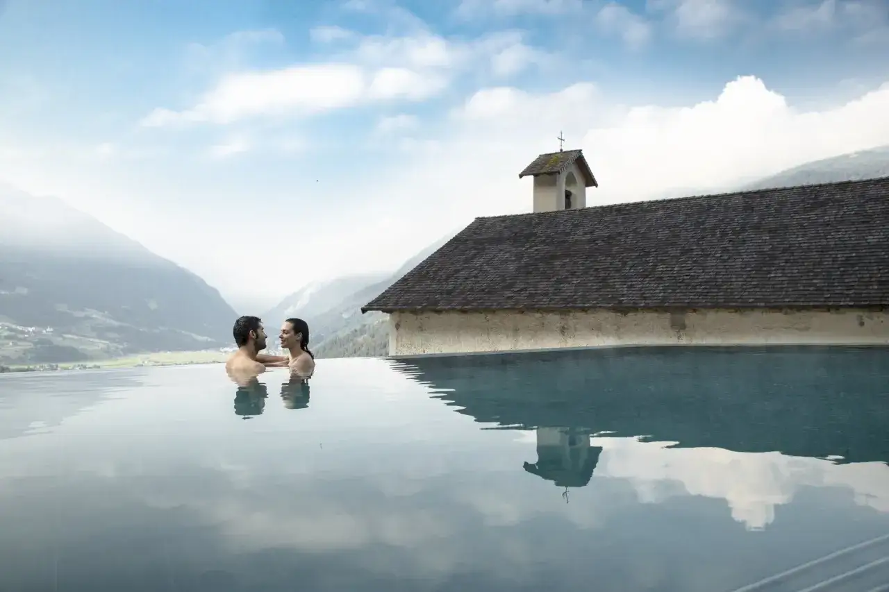 Coppia in piscina a sfioro con vista sulle montagne di Bormio e dintorni. Riflessi sull'acqua e un antico edificio con campanile.