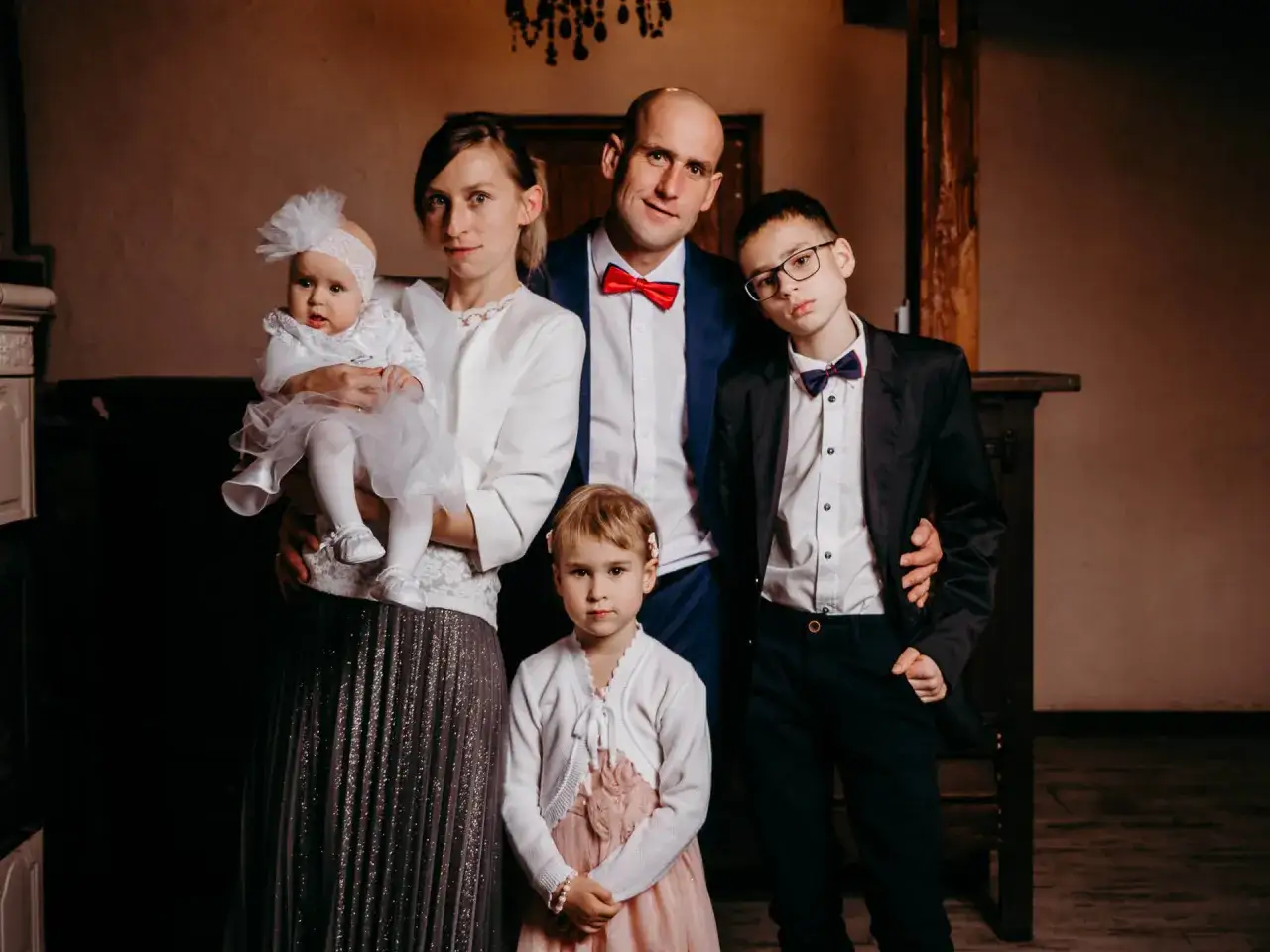 Rodzina świętuje chrzest dziecka. Mama tuli niemowlę w białej sukience, obok stoją tata i dwójka starszych dzieci.