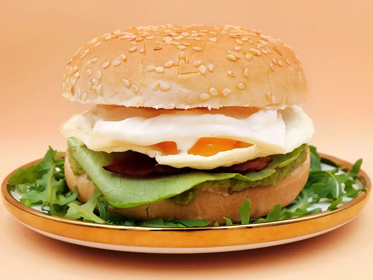 Pyszny burger z jajkiem sadzonym, chrupiącym boczkiem i świeżą rukolą na złotej talerzy.
