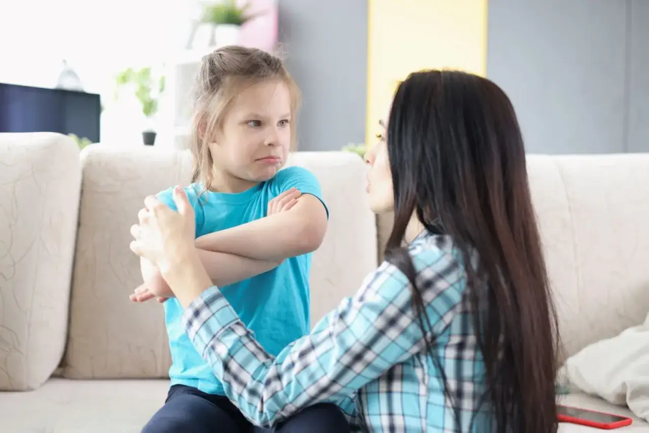 Bambina di 10 anni con espressione accigliata e braccia incrociate, mentre una donna cerca di calmarla. Potrebbe essere una crisi di rabbia.