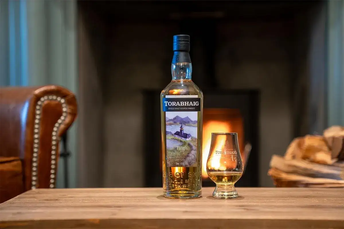 Torabhaig peated whisky in einem Glas, daneben die Flasche, vor einem Kaminfeuer und einem Ledersessel.