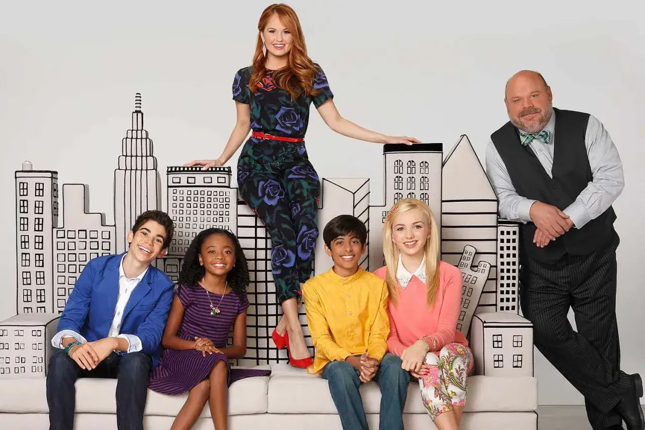 Jessie program telewizyjny obsada: Debby Ryan, Cameron Boyce, Skai Jackson, Karan Brar, Peyton List i Charles Esten pozują na tle rysunkowego miasta.