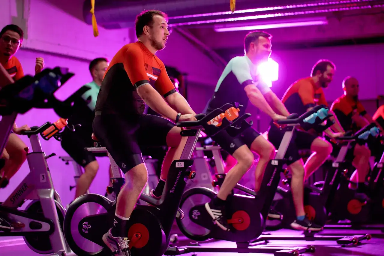 Grupa osób ćwiczy na rowerach stacjonarnych w sali o fioletowym oświetleniu. To właśnie indoor cycling – intensywny trening kolarski w pomieszczeniu.