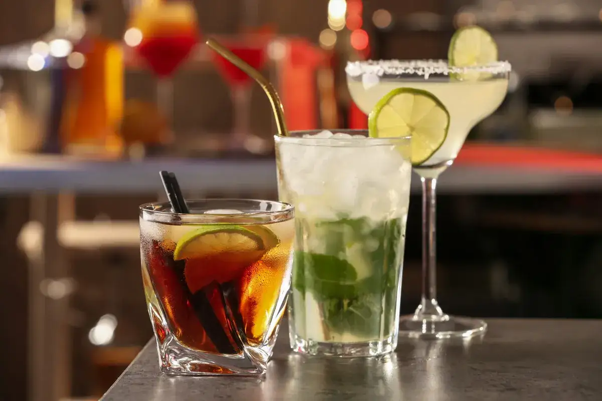 Tre läckra drinkar med rom, lime och mynta står på en bardisk. En mörk romdrink med is och lime, en mojito och en ljusare drink i ett martiniglas.