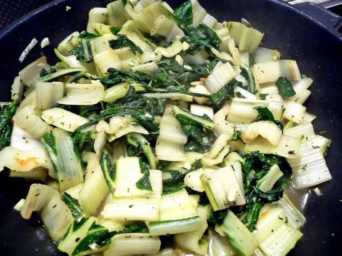 Cuisinant des côtes de blettes tendres et vertes dans une poêle, assaisonnées d'herbes.