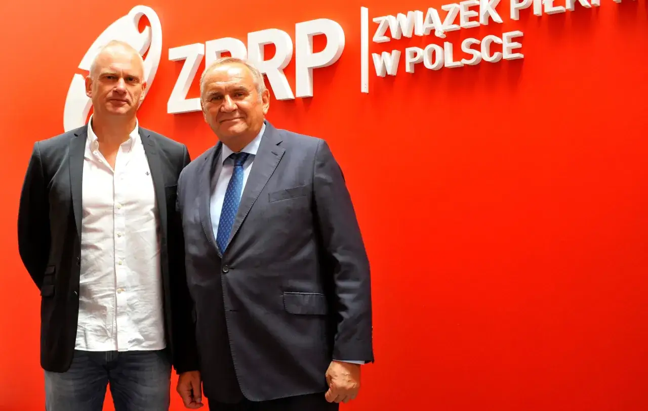 Dw&oacute;ch mężczyzn na tle czerwonej ściany z logo ZPRP. Jeden z nich to trener reprezentacji Polski w piłce ręcznej.