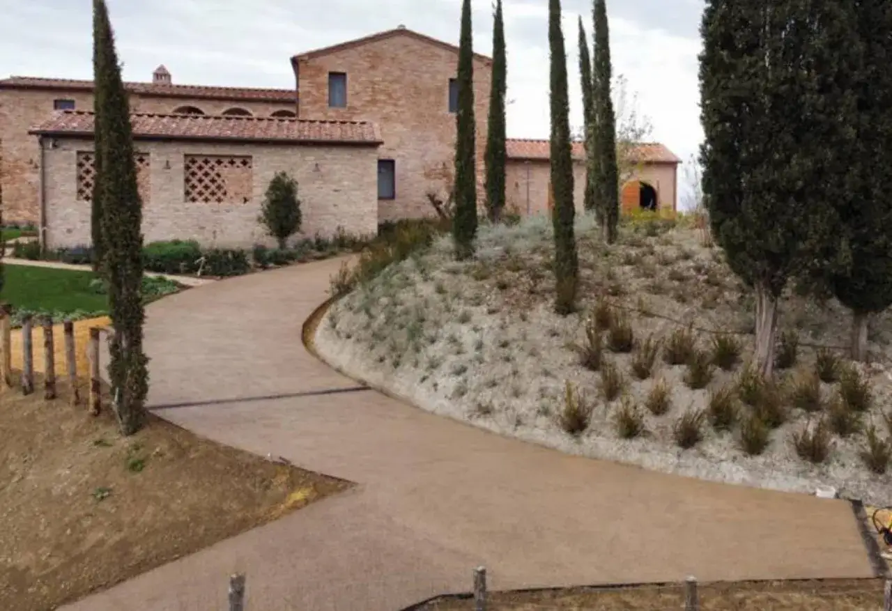 Strada in asfalto permeabile che conduce a un edificio in mattoni con cipressi e vegetazione mediterranea.