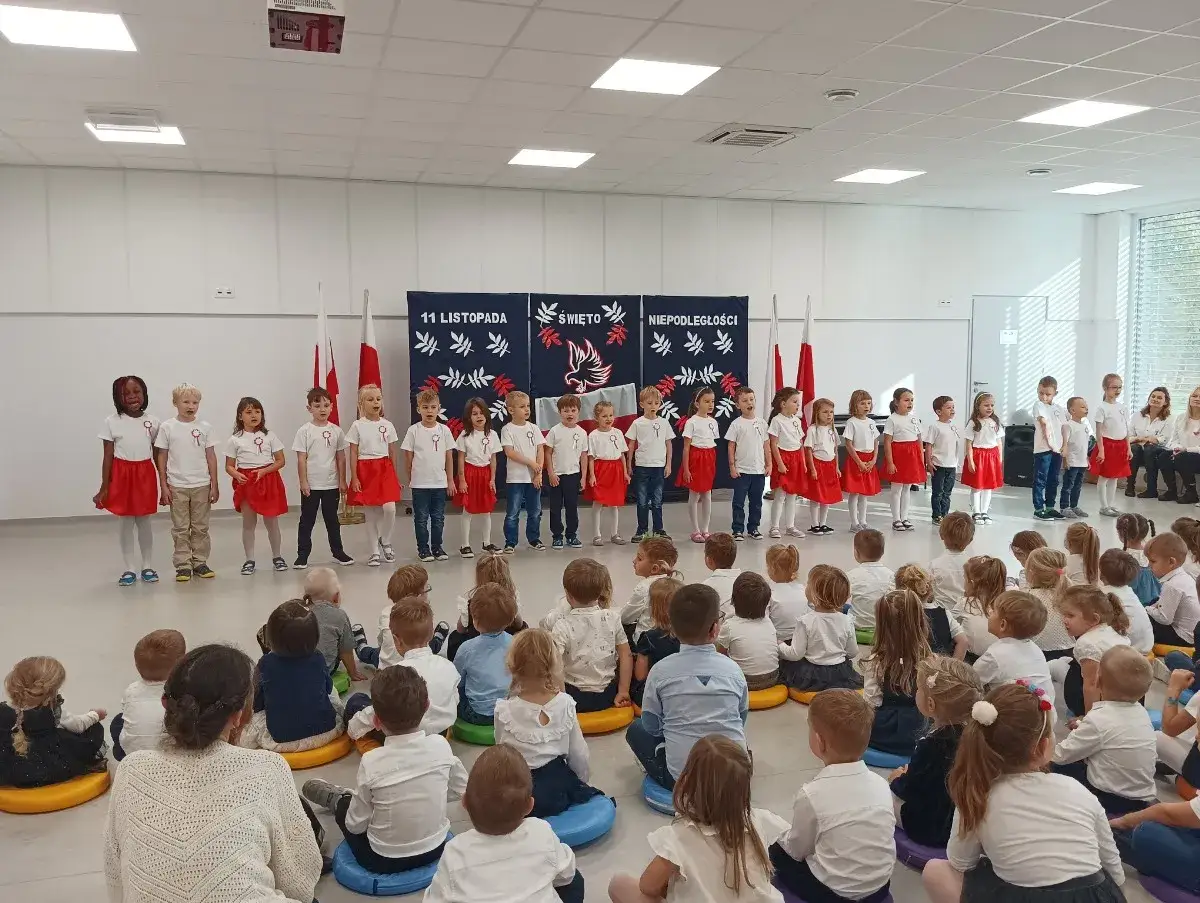 Dzieci w białych koszulkach i czerwonych spódniczkach śpiewają piosenki patriotyczne na 11 listopada.